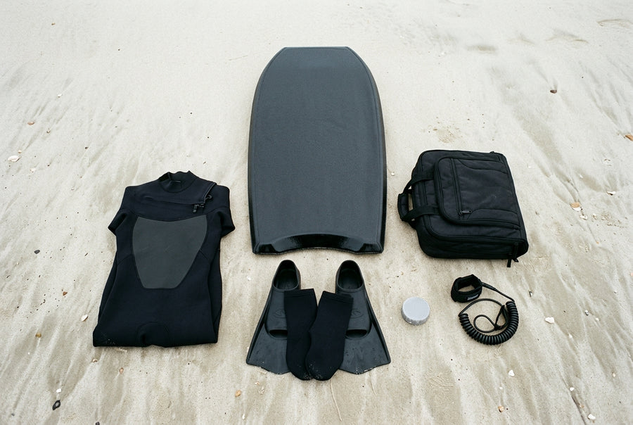 Equipo completo del bodyboarder sobre la arena: tabla, aletas, leash, traje de neopreno, calcetines, wax y funda