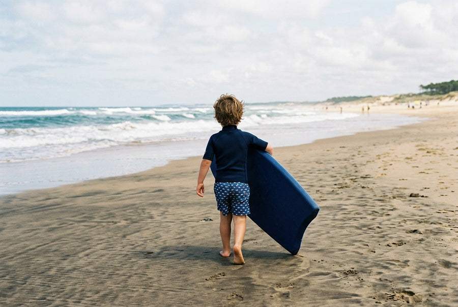 Jeune rider de dos portant une planche de bodyboard trop grande pour lui vers l'eau